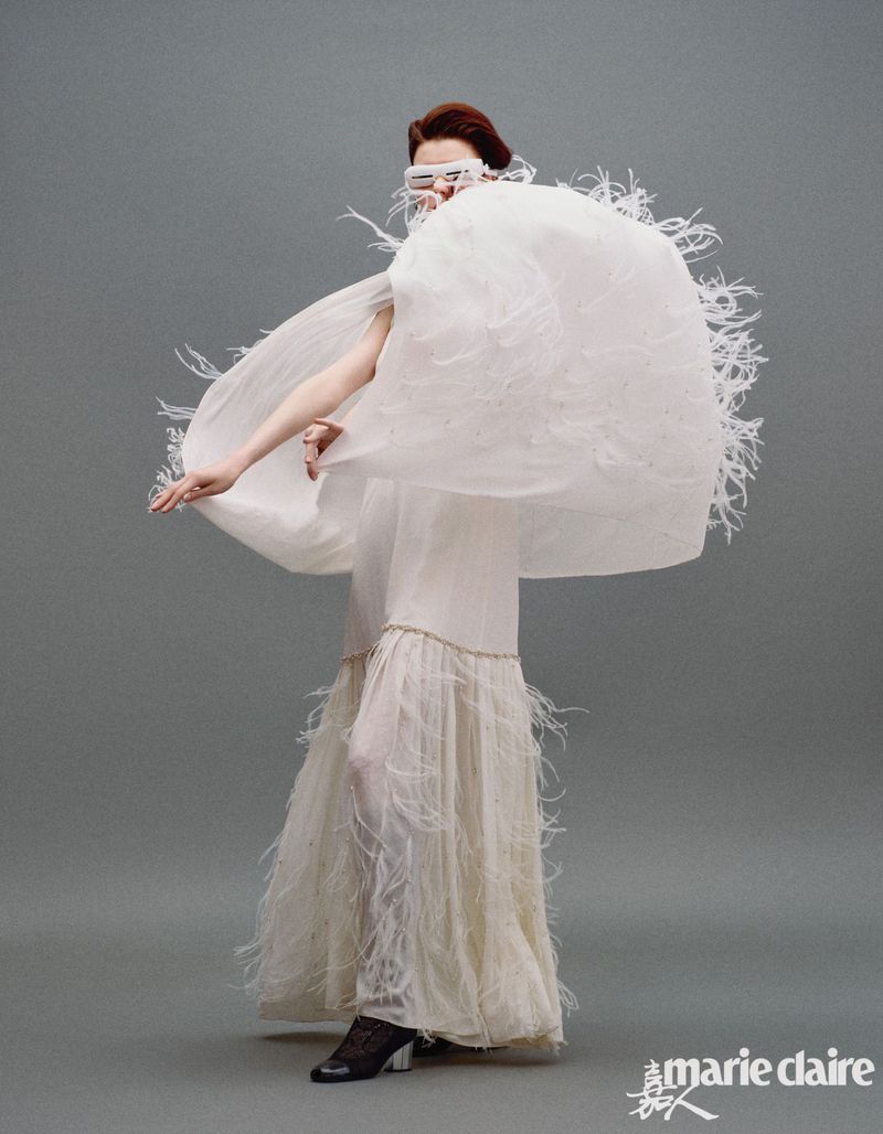 A stylish woman in a white, feathered gown and futuristic glasses strikes a pose against a neutral grey backdrop for a fashion editorial.