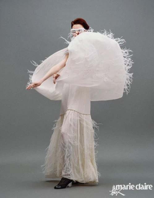 A stylish woman in a white, feathered gown and futuristic glasses strikes a pose against a neutral grey backdrop for a fashion editorial.