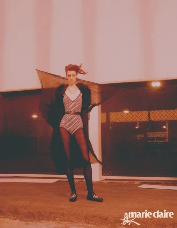 A woman stands in a patterned bodysuit and black tights, wearing a fur-trimmed wrap, posing outdoors in front of a commercial building at night.