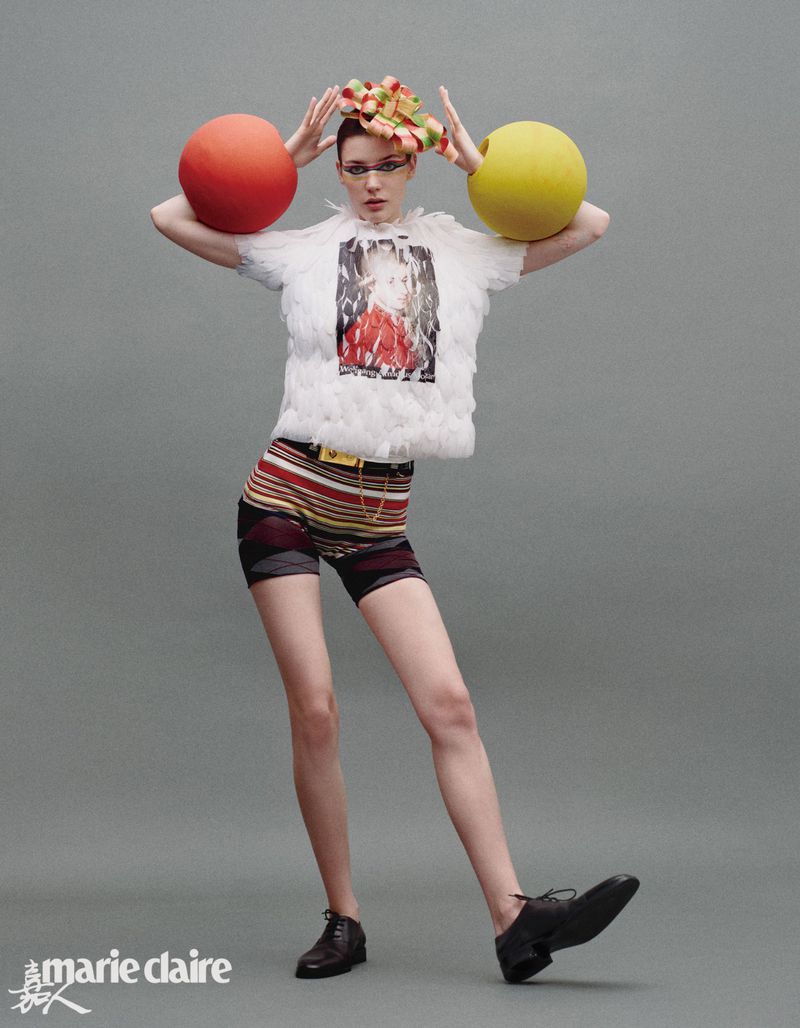 A woman poses in a white feathered top and striped shorts with large colored balls on either side of her head in a studio shot.
