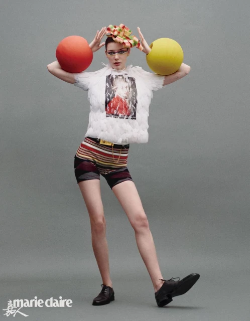A woman poses in a white feathered top and striped shorts with large colored balls on either side of her head in a studio shot.