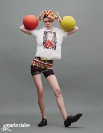 A woman poses in a white feathered top and striped shorts with large colored balls on either side of her head in a studio shot.