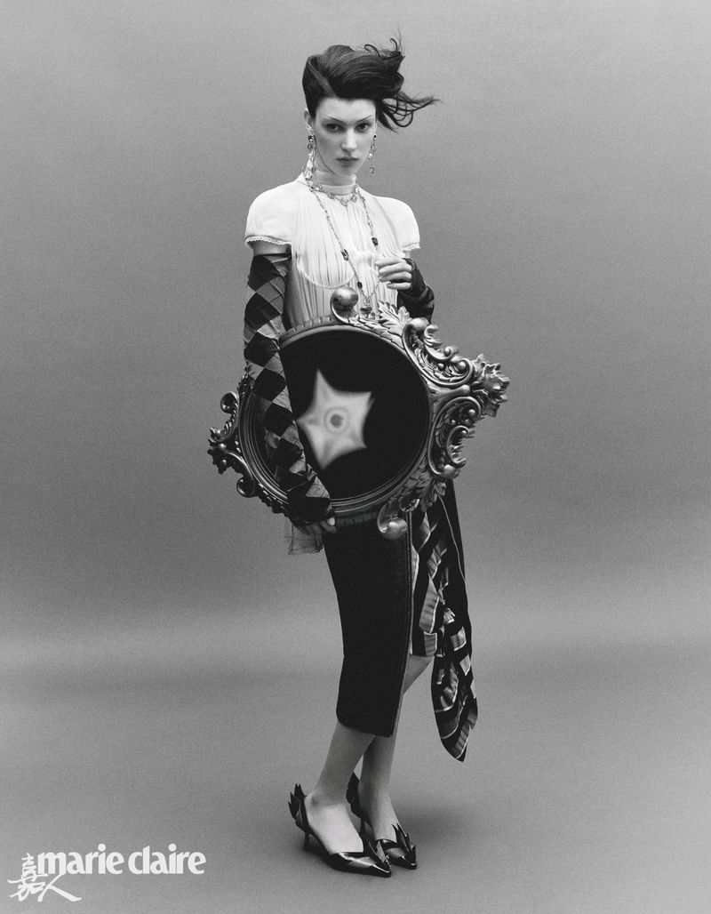 Black and white fashion portrait of a woman with artistic makeup, sculptural hair, and patterned dress holding an ornate mirror.