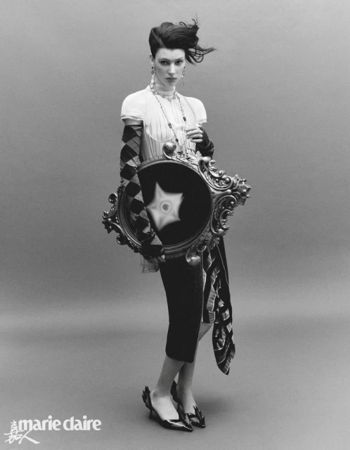 Black and white fashion portrait of a woman with artistic makeup, sculptural hair, and patterned dress holding an ornate mirror.