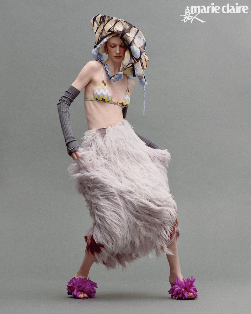 A model strikes a pose in a studio setting, showcasing a bikini top, fur skirt, floral platform sandals, and a unique headpiece in a fashion editorial.