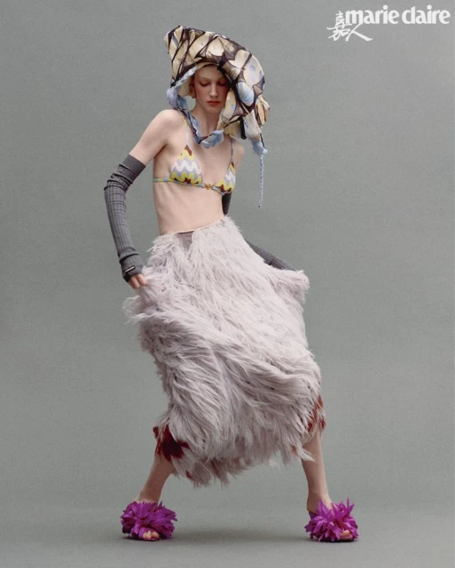 A model strikes a pose in a studio setting, showcasing a bikini top, fur skirt, floral platform sandals, and a unique headpiece in a fashion editorial.