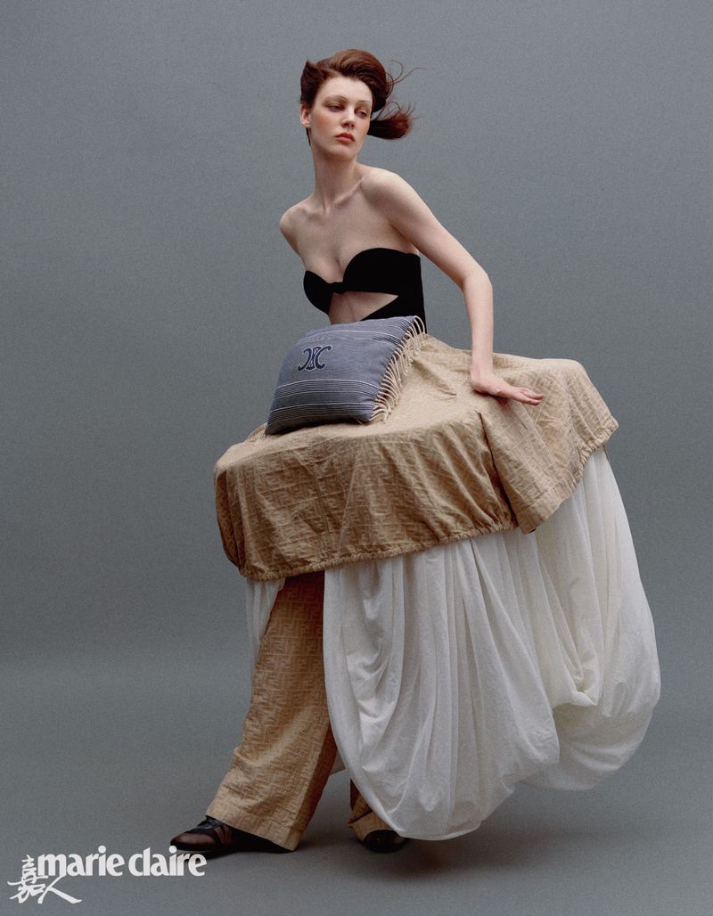 Editorial fashion shot of a woman in couture, featuring a strapless top, pillow, beige table skirt, and matching pants against a gray backdrop.