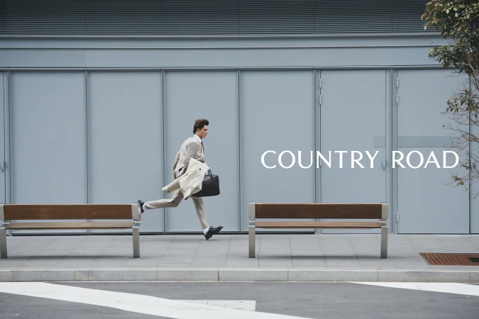 A man in a suit runs in an urban setting, carrying a coat and bag near benches and a grey building, showing movement and urgency.