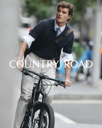 A young man in smart casual clothing rides a bicycle on a city street, with the Country Road logo prominently displayed.