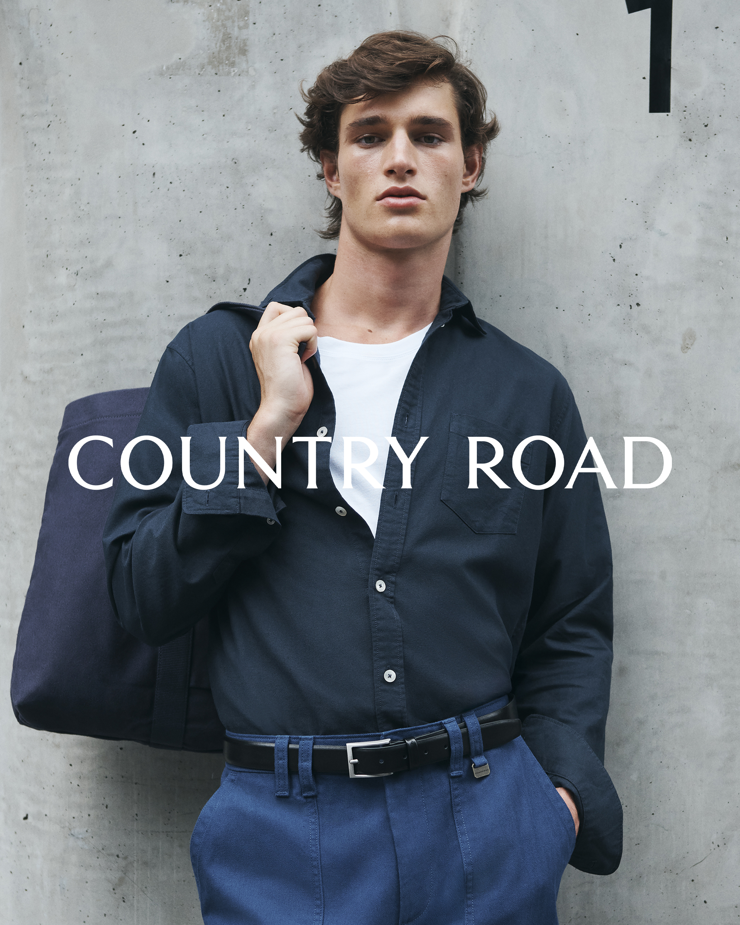 A young man in a casual blue outfit with a bag slung over his shoulder, posing against a concrete wall with the brand Country Road.