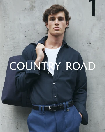 A young man in a casual blue outfit with a bag slung over his shoulder, posing against a concrete wall with the brand Country Road.