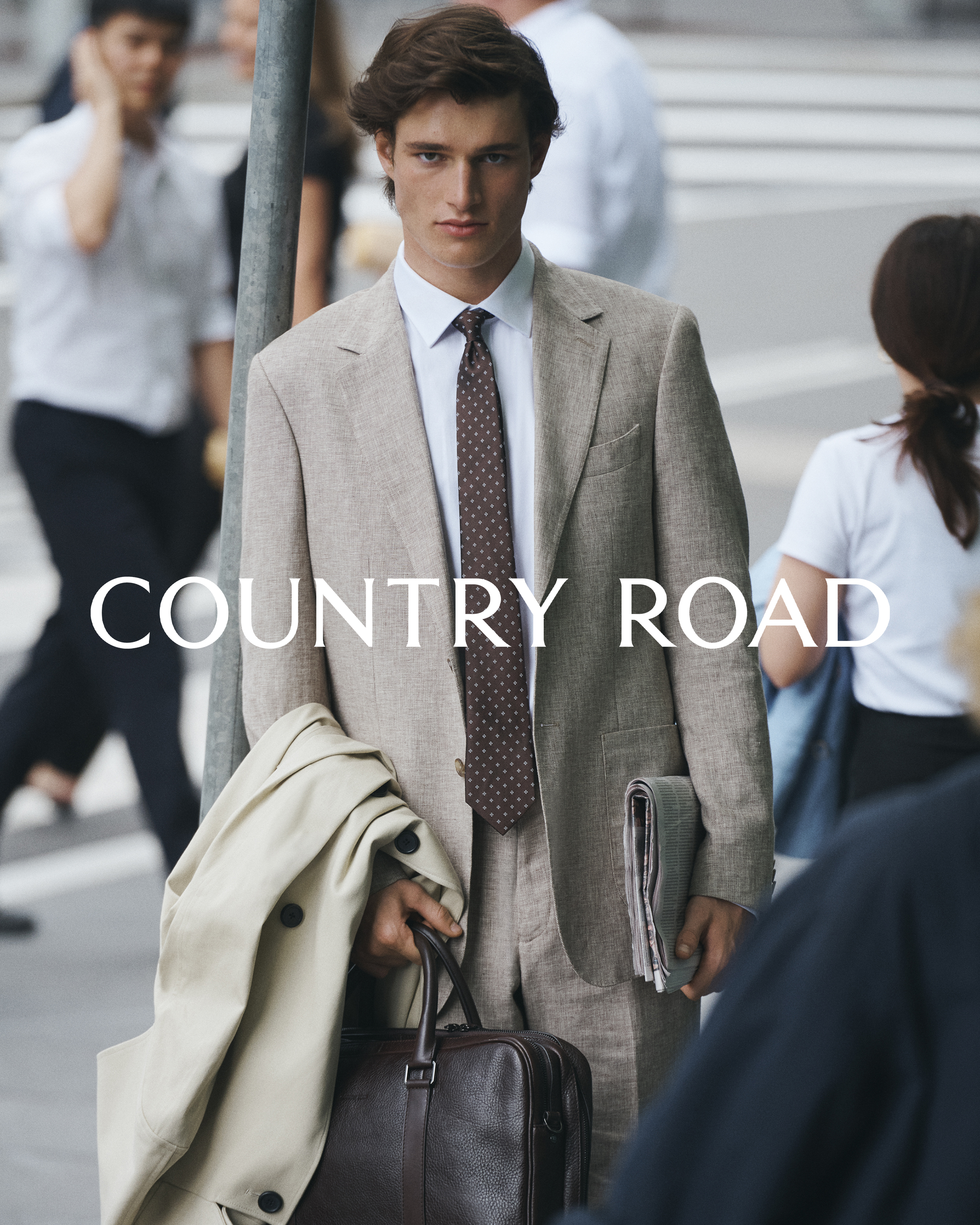 A man in a suit and tie stands on a city street, holding a briefcase and a coat, with the text 'COUNTRY ROAD' visible.