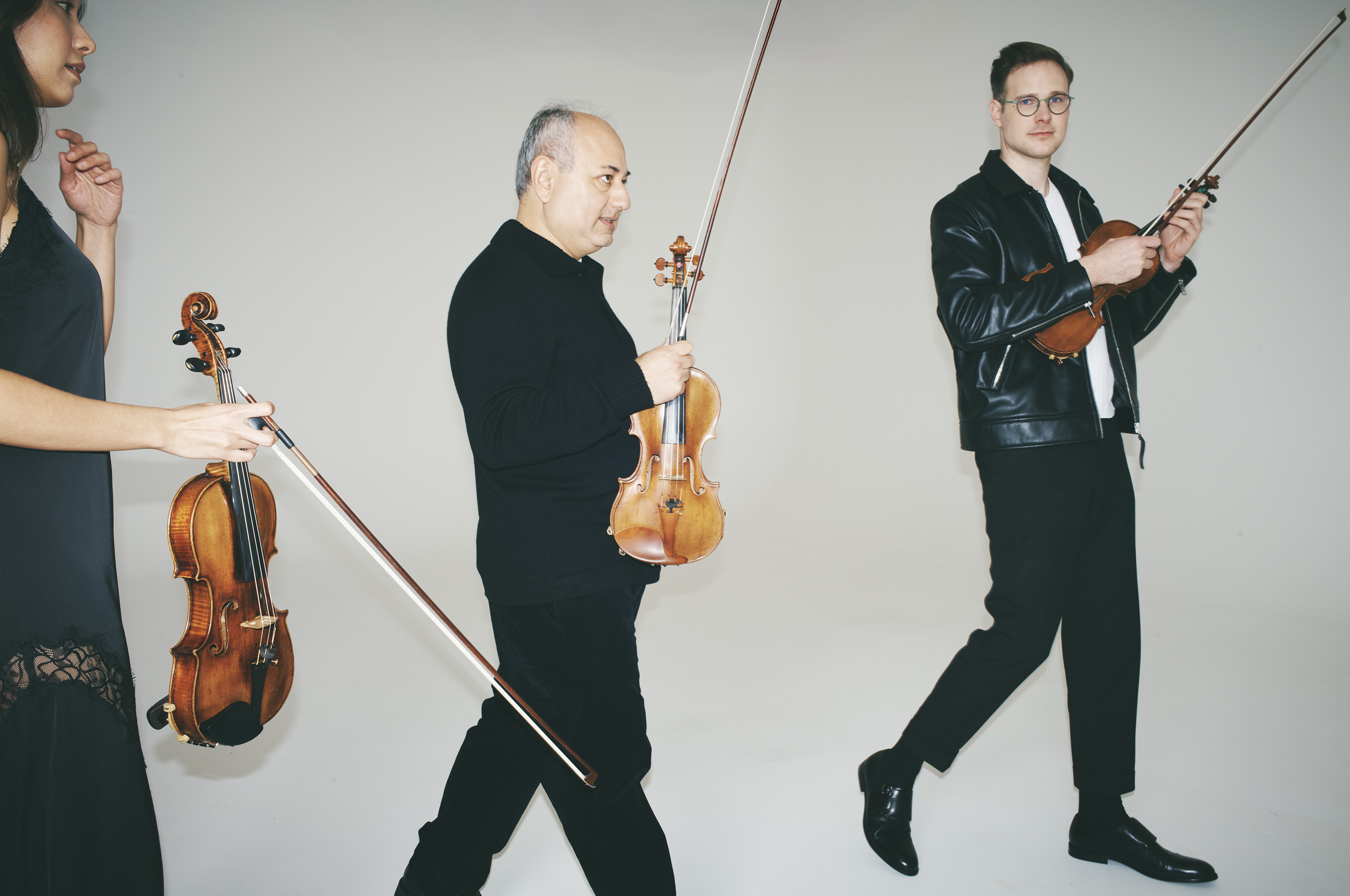 Three musicians walk carrying violins and bows in a neutral indoor studio. One woman and two men, all in dark clothing, hold their instruments.