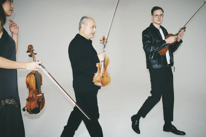Three musicians walk carrying violins and bows in a neutral indoor studio. One woman and two men, all in dark clothing, hold their instruments.