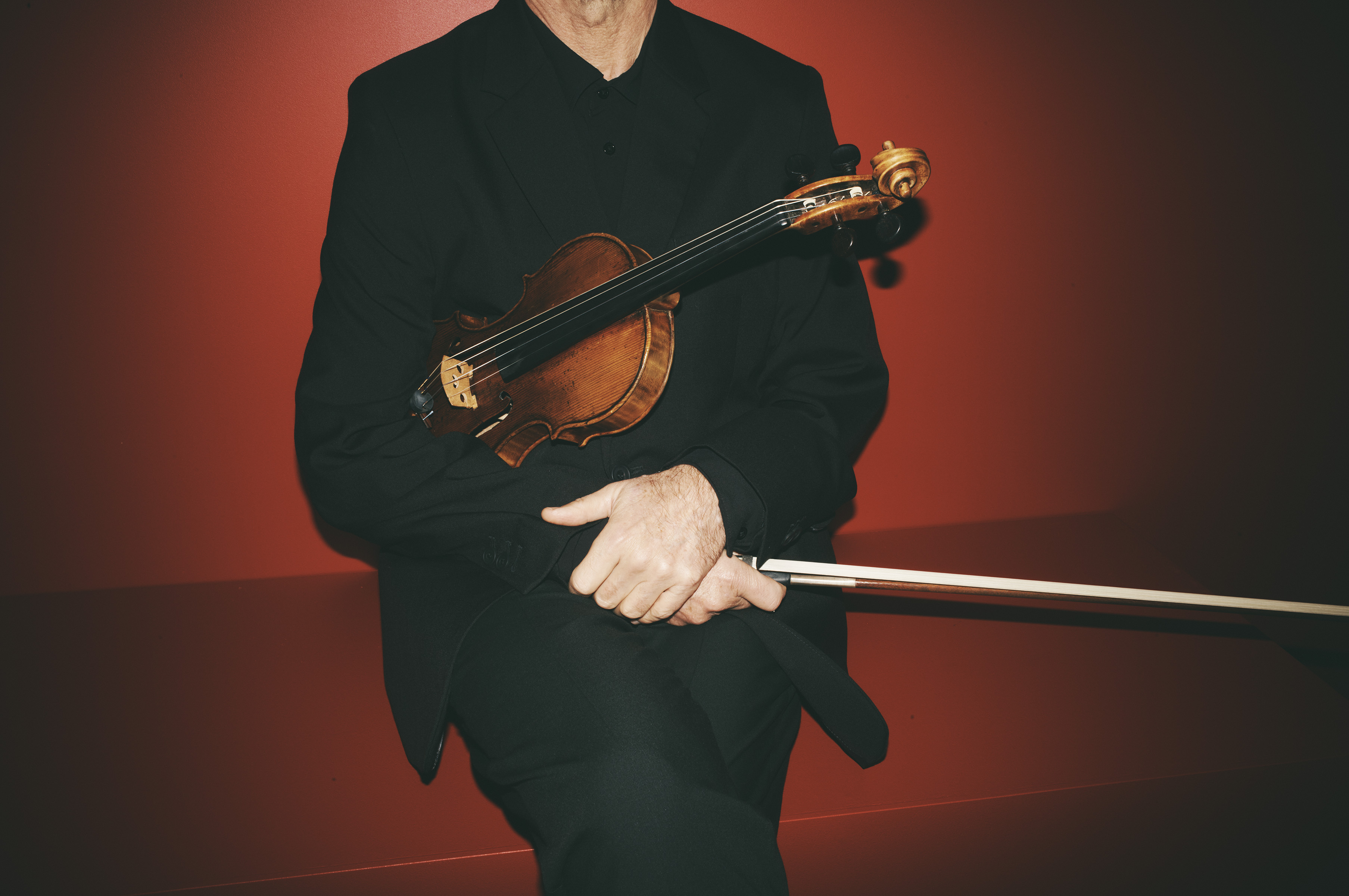 A man in a black suit holds a violin and bow in front of a red background, creating a contrast between the subject and backdrop.