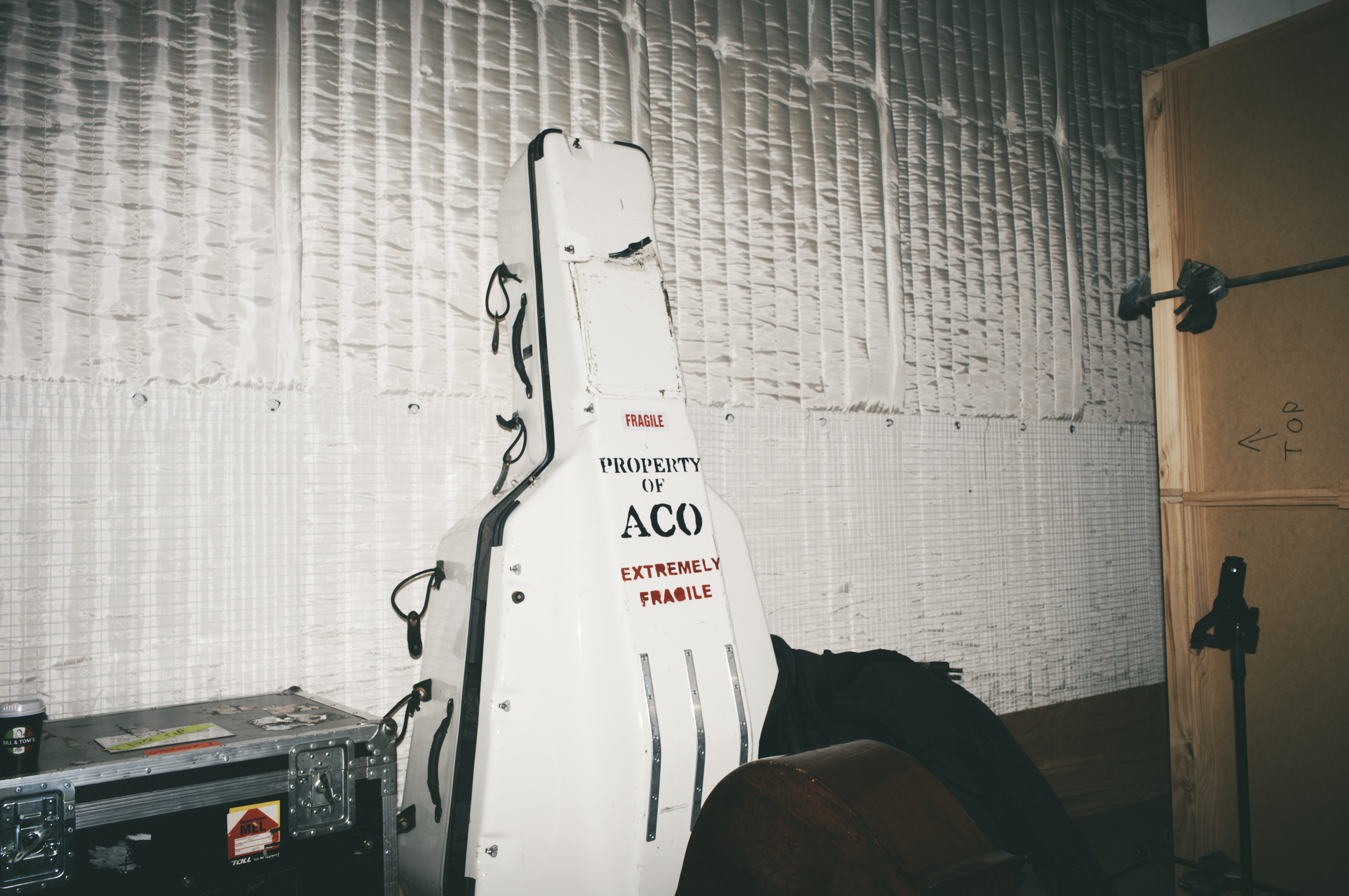 Indoor view of a guitar case with 'FRAGILE' and 'PROPERTY OF ACO' printed on it, sitting next to road cases and a wooden panel.