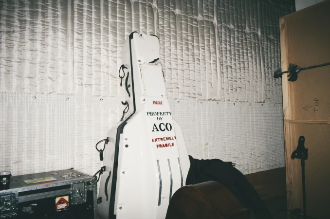 Indoor view of a guitar case with 'FRAGILE' and 'PROPERTY OF ACO' printed on it, sitting next to road cases and a wooden panel.