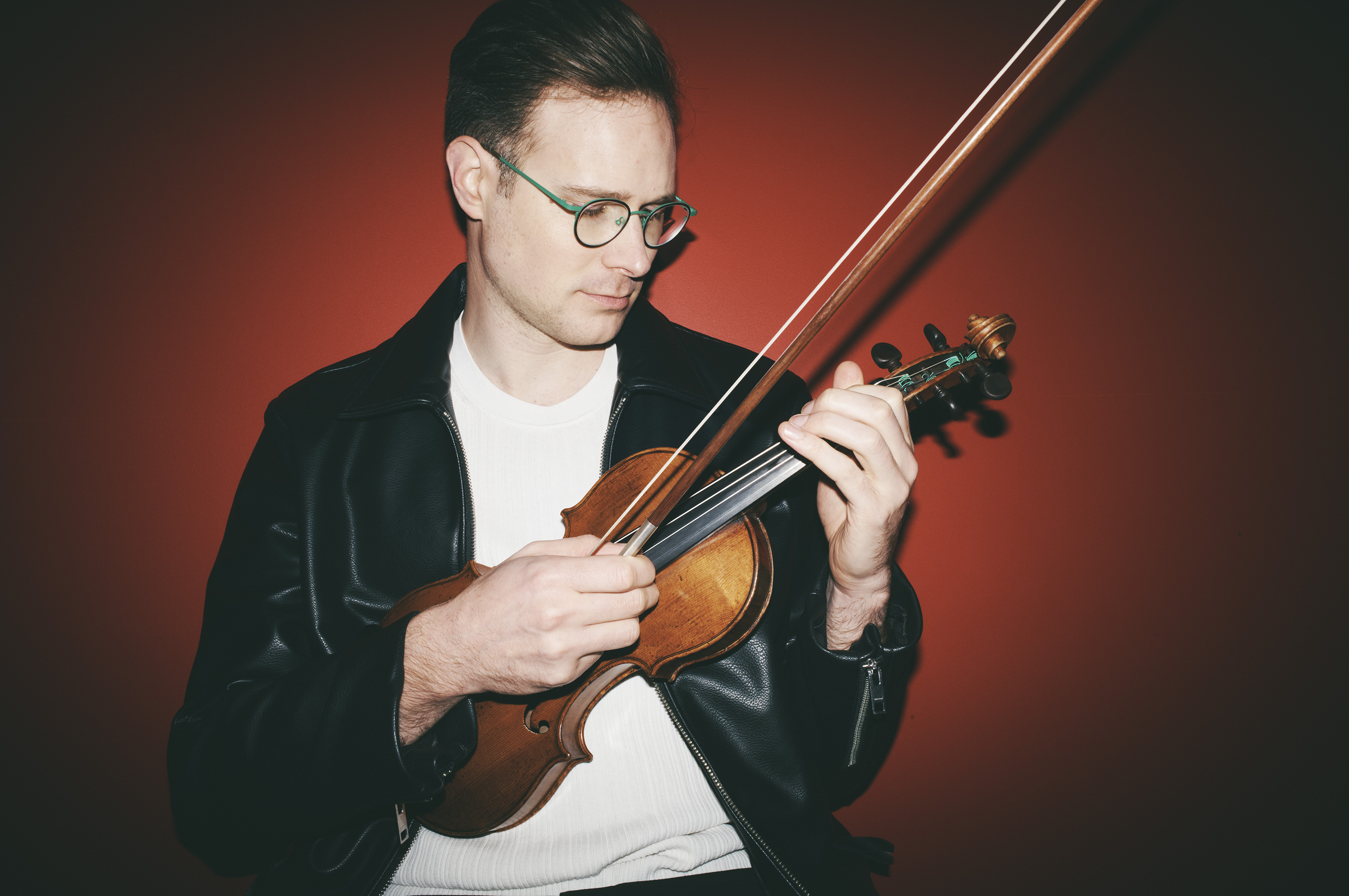 A man with glasses and a black leather jacket plays the violin against a red backdrop, showcasing music and style in a close-up shot.