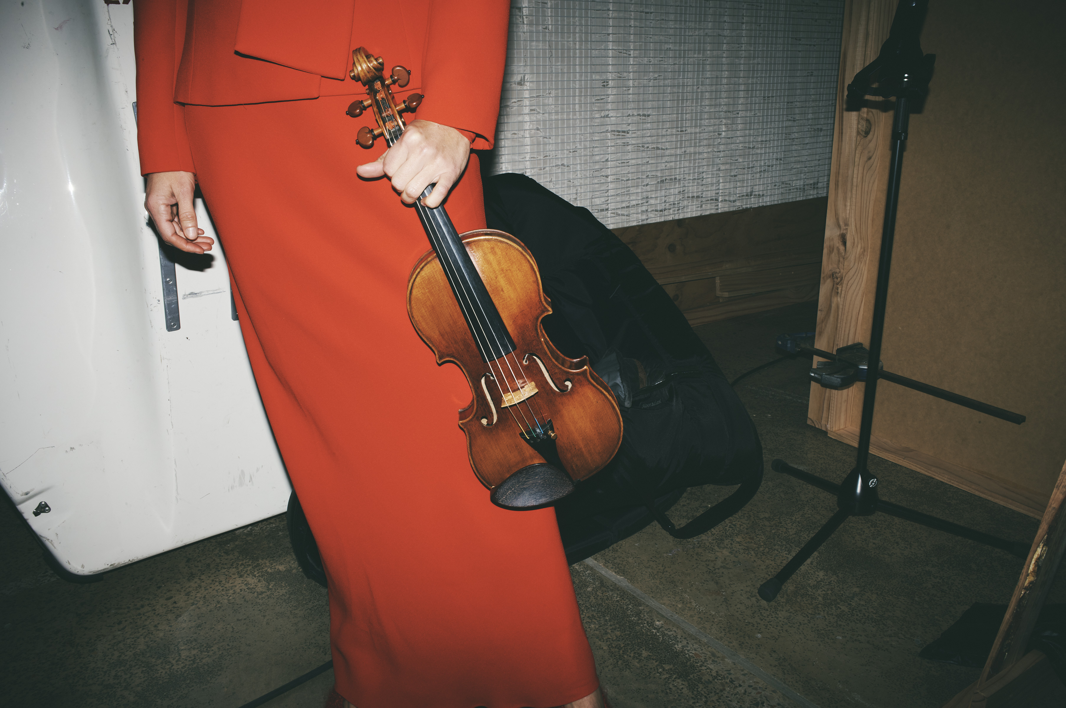 Woman in red dress holding a violin indoors, with black case and microphone stand in the background, low angle shot, grainy texture.