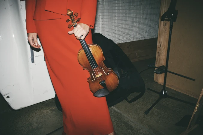 Woman in red dress holding a violin indoors, with black case and microphone stand in the background, low angle shot, grainy texture.
