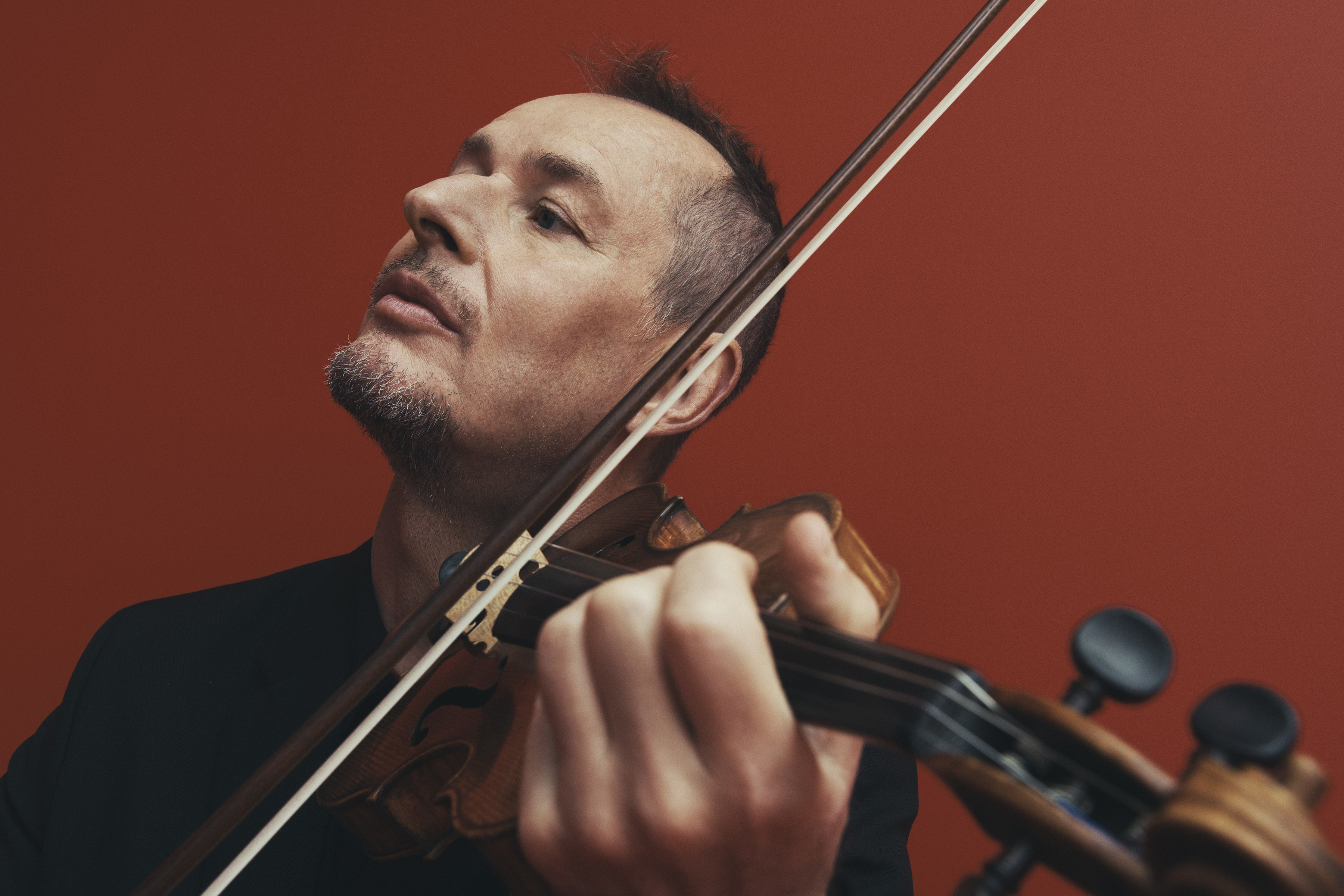 Man plays the violin against an orange background. The musician is wearing a dark suit jacket and the focus is on his face and instrument.