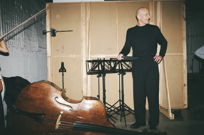 Indoor shot of a male conductor standing near a double bass with a music stand in the background, in a room with panelled walls.