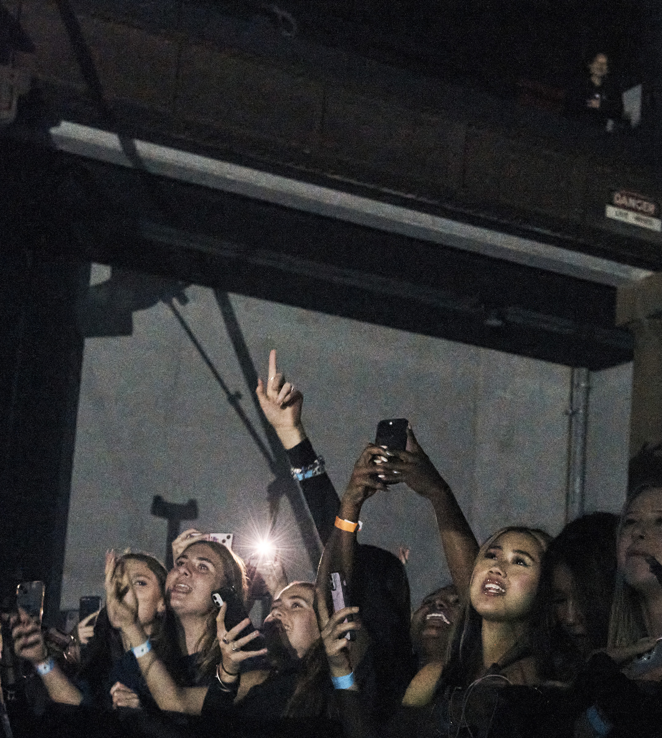 Large crowd of diverse people holding up phones and looking up in a dimly lit indoor venue, likely at a concert or music event.