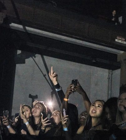 Large crowd of diverse people holding up phones and looking up in a dimly lit indoor venue, likely at a concert or music event.