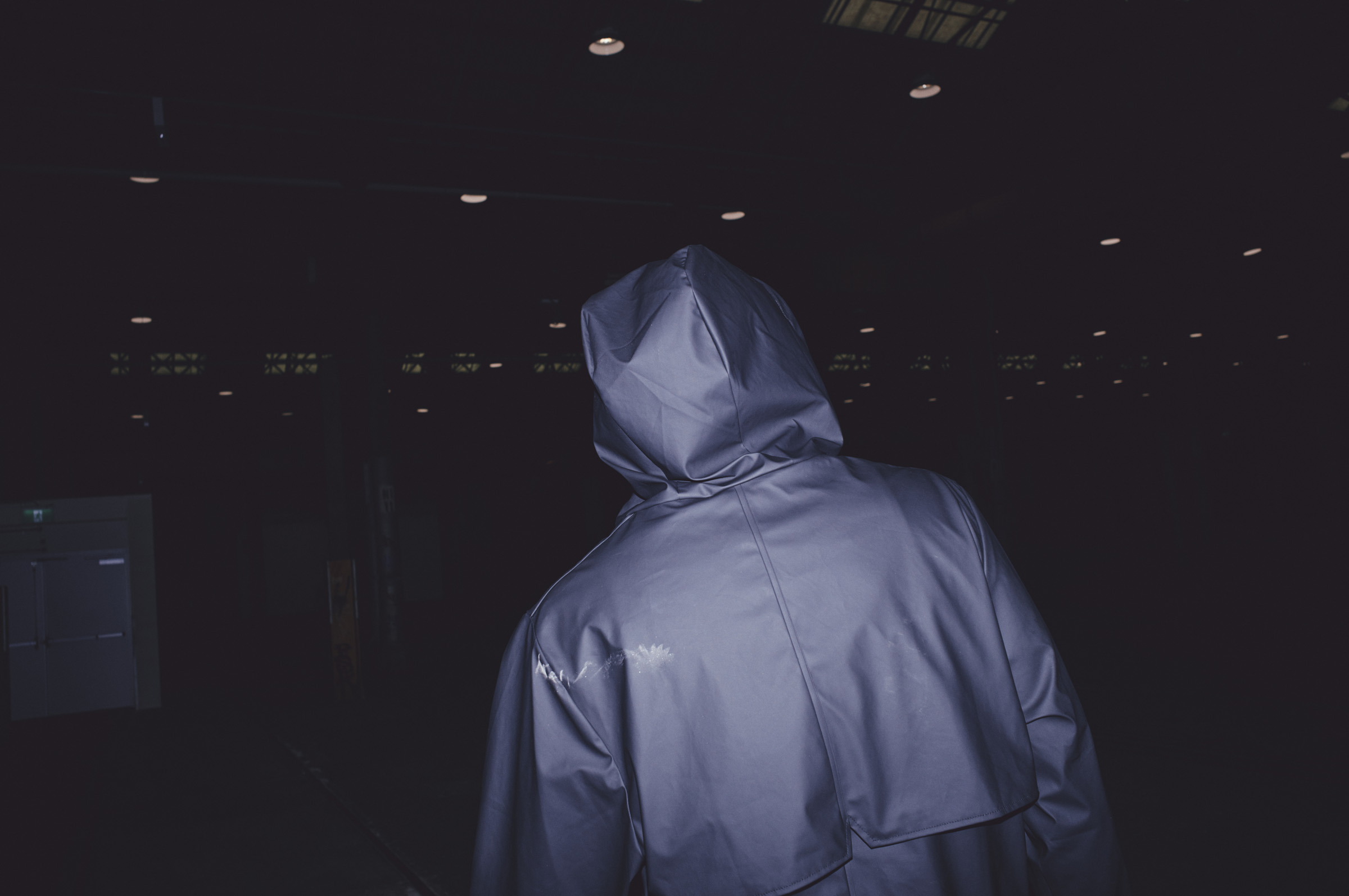 Person in a hooded blue jacket walks through a dimly lit building interior, back to the camera, anonymous and isolated figure.