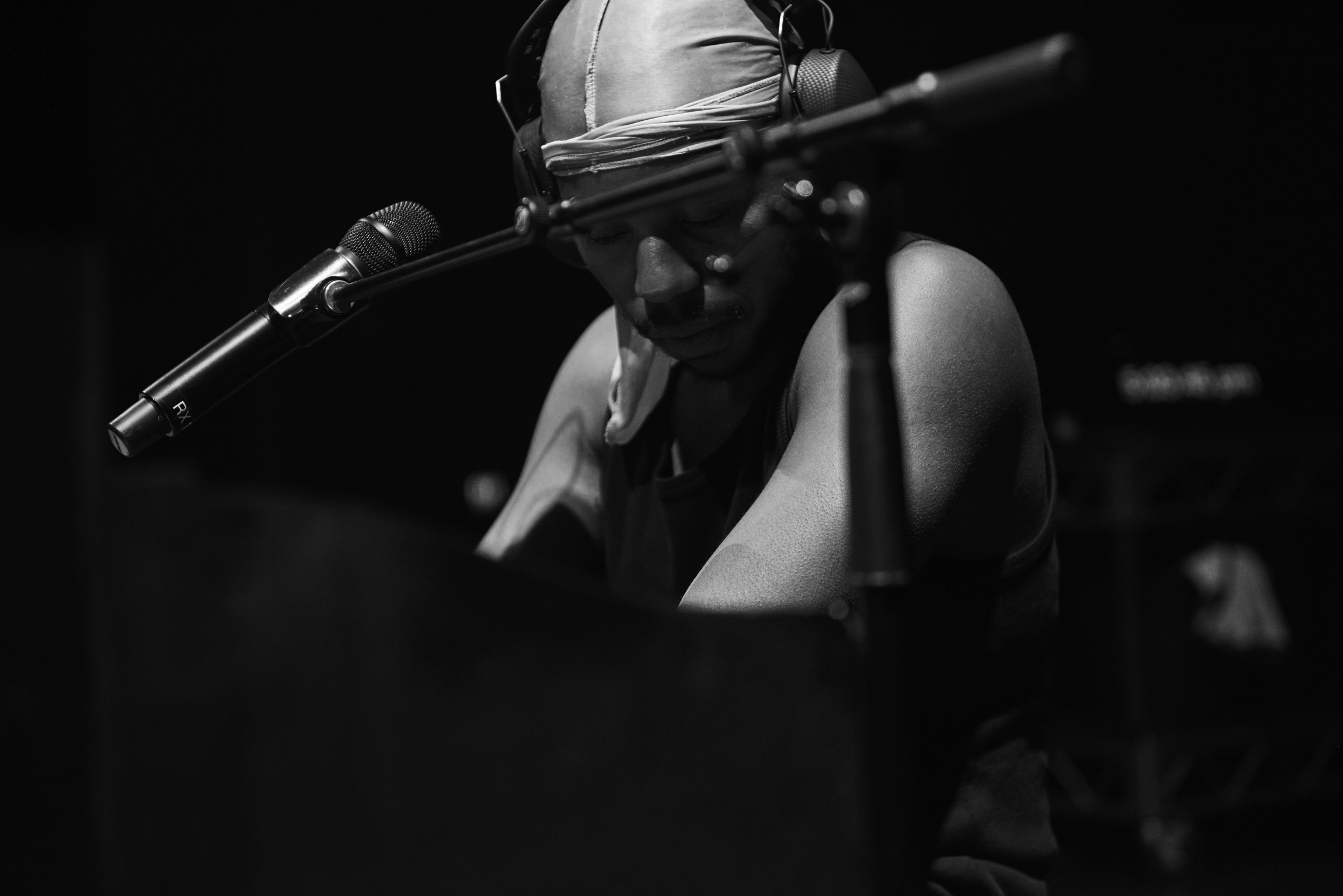 Black and white image of a musician wearing headphones and head covering playing a keyboard with a microphone in a dark setting.