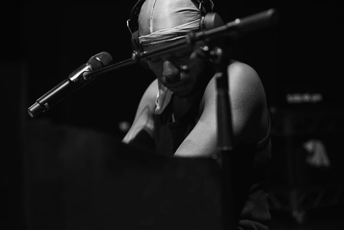 Black and white image of a musician wearing headphones and head covering playing a keyboard with a microphone in a dark setting.