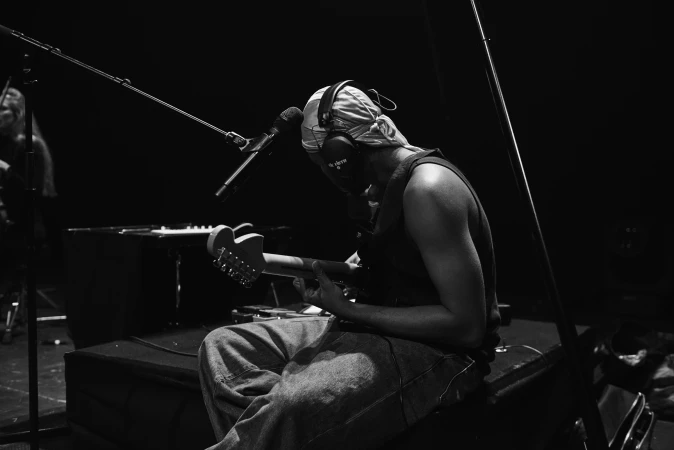 Monochrome shot of a musician in headphones playing electric guitar, seated in a dark music recording studio with microphones and keyboard.