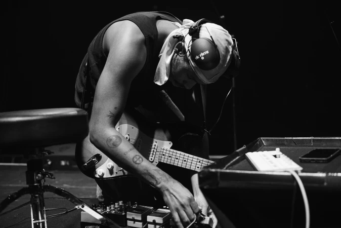 Monochrome shot of musician with head covering and headphones, playing guitar and adjusting controls on a synthesizer onstage.