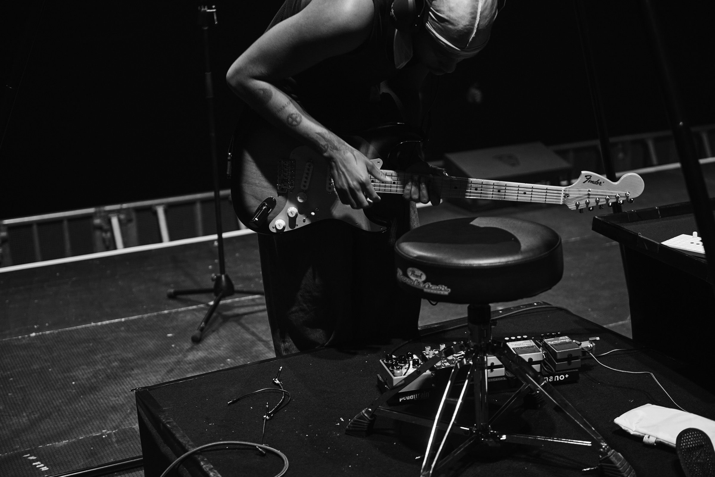 Black and white close-up of a musician playing an electric guitar on stage, seated on a stool with musical equipment nearby.