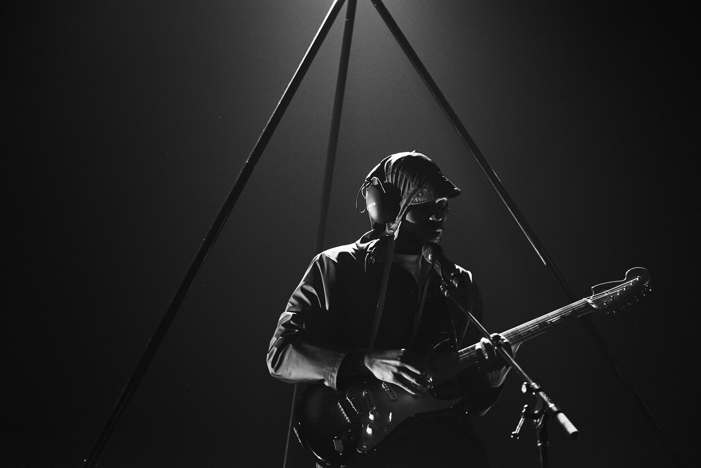 Monochrome portrait of a musician with a head covering, playing an electric guitar on stage with a geometric frame and dark background.