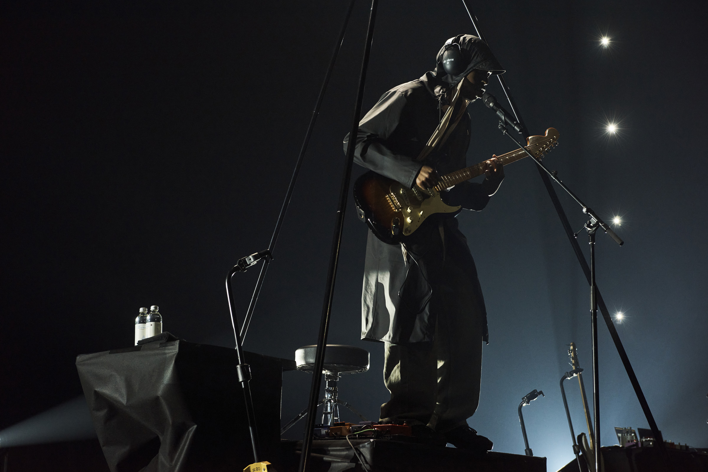 Musician performs on stage with guitar, wearing unique headgear and jacket, on elevated platform with microphones and equipment.