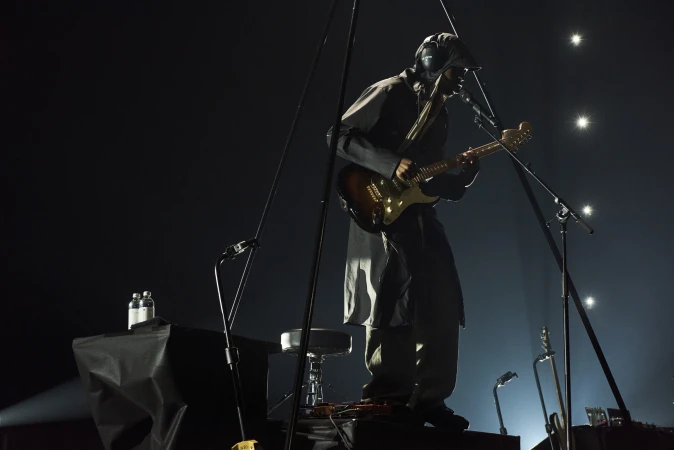 Musician performs on stage with guitar, wearing unique headgear and jacket, on elevated platform with microphones and equipment.