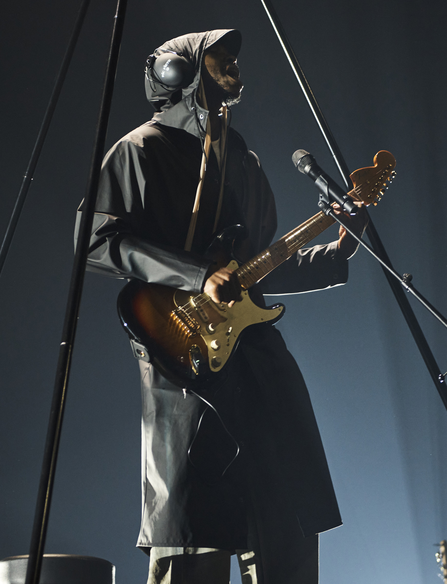 Musician in hooded coat performs with electric guitar and microphone on stage with rigging and dark background, bathed in soft light.