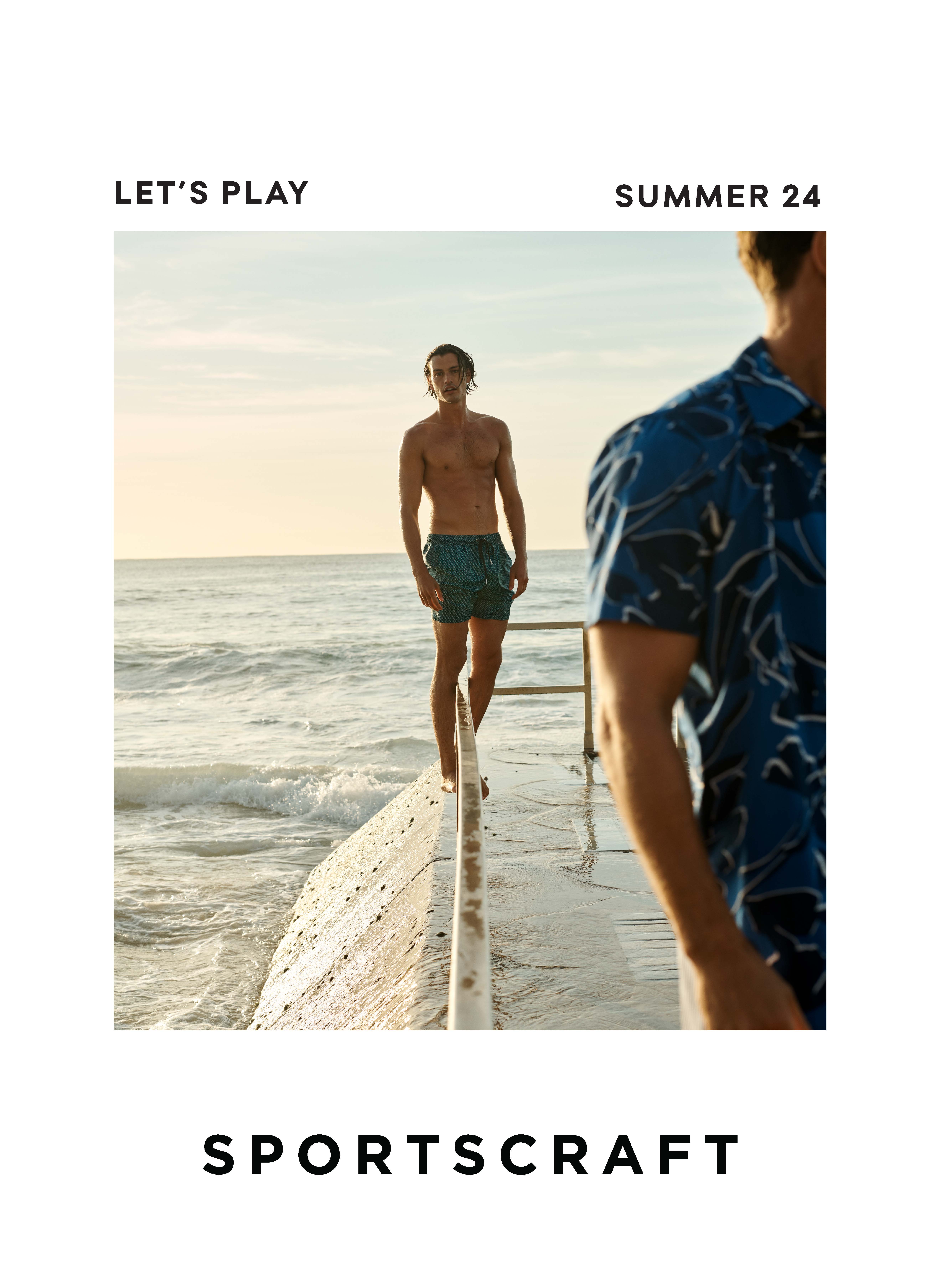 Two adult men pose on a concrete structure with ocean in background during daytime, wearing swimwear and button-down shirt.