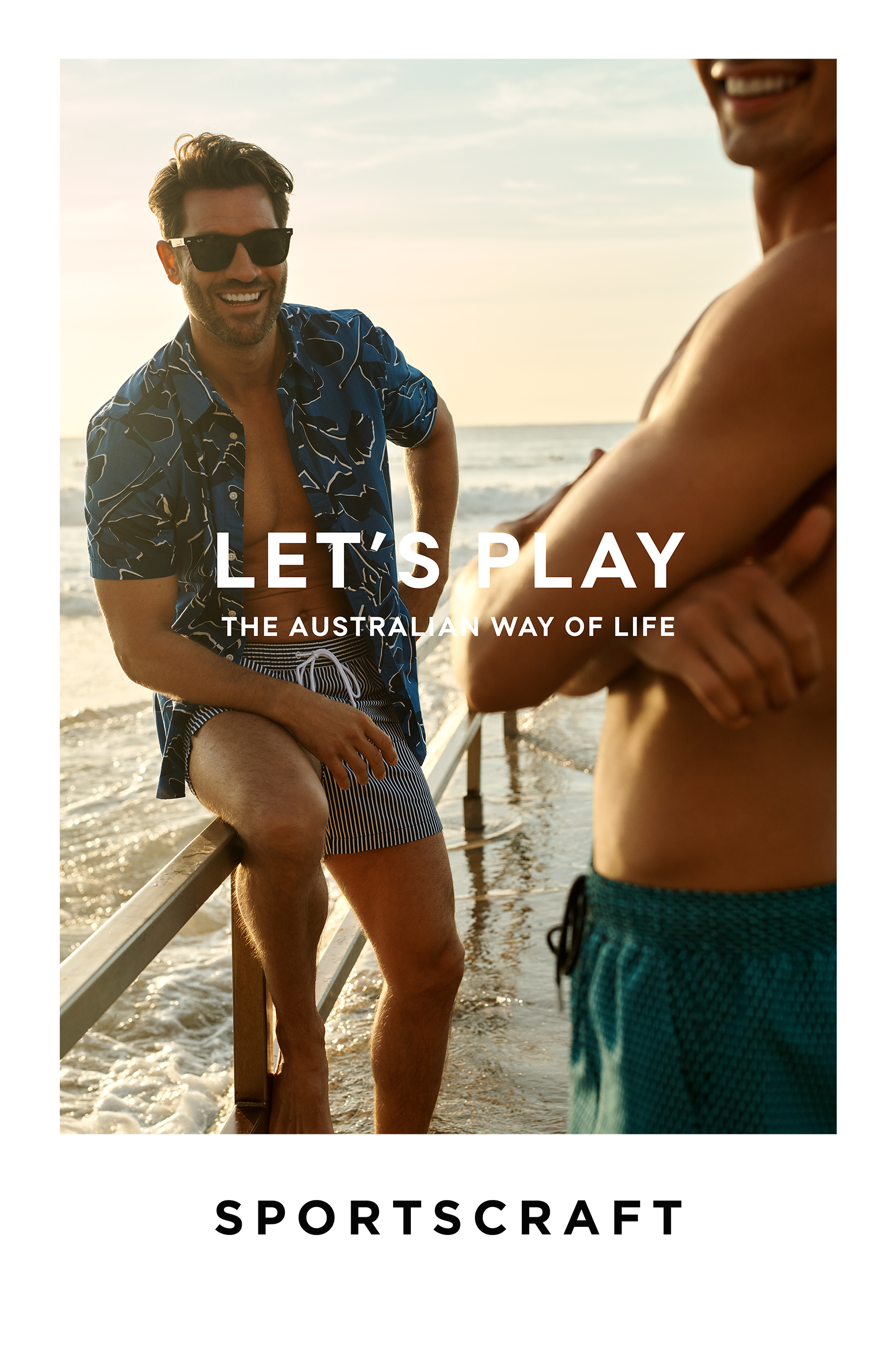 Two adult men on a beach with ocean waves and text overlay, in swimwear and casual open shirts, enjoying a summer day.
