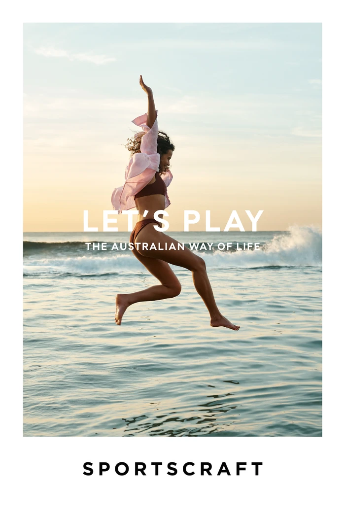 Woman mid-air leaping over ocean waves at sunset wearing a pink shirt and brown bikini, photographed by Simon Lekias.