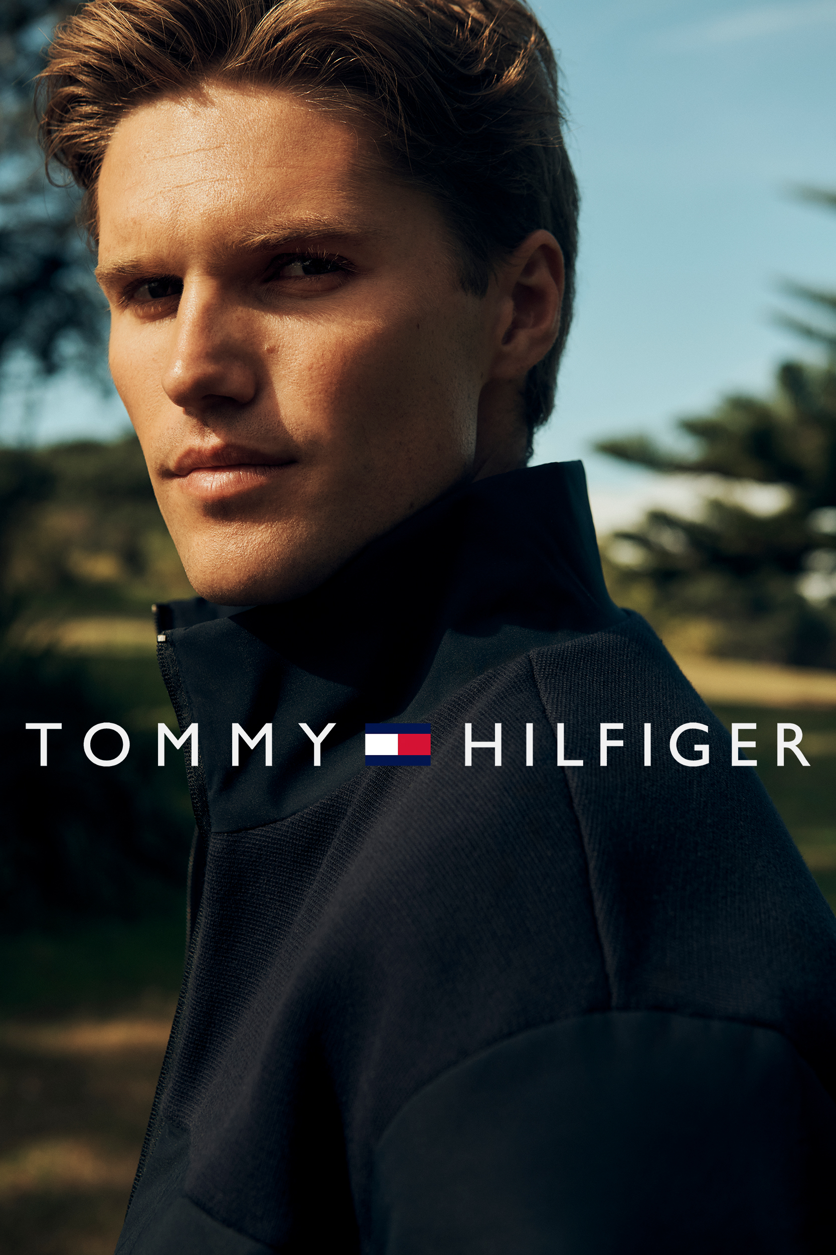 Close-up shot of a blond male model wearing a navy blue jacket, showcasing Tommy Hilfiger logo against a soft outdoor background.