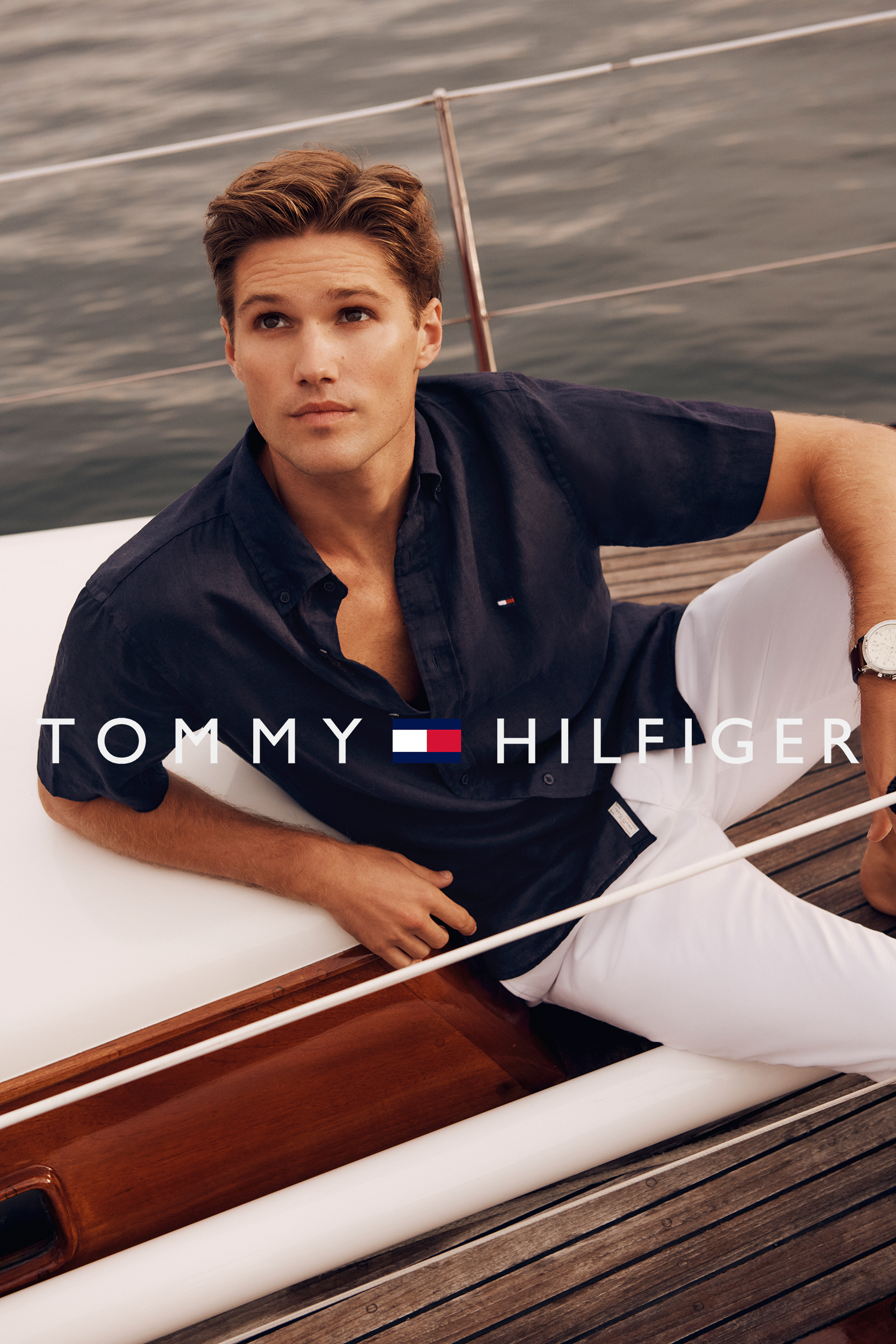Handsome male model in navy shirt and white pants lounges on a yacht, promoting Tommy Hilfiger in a nautical-themed fashion campaign.