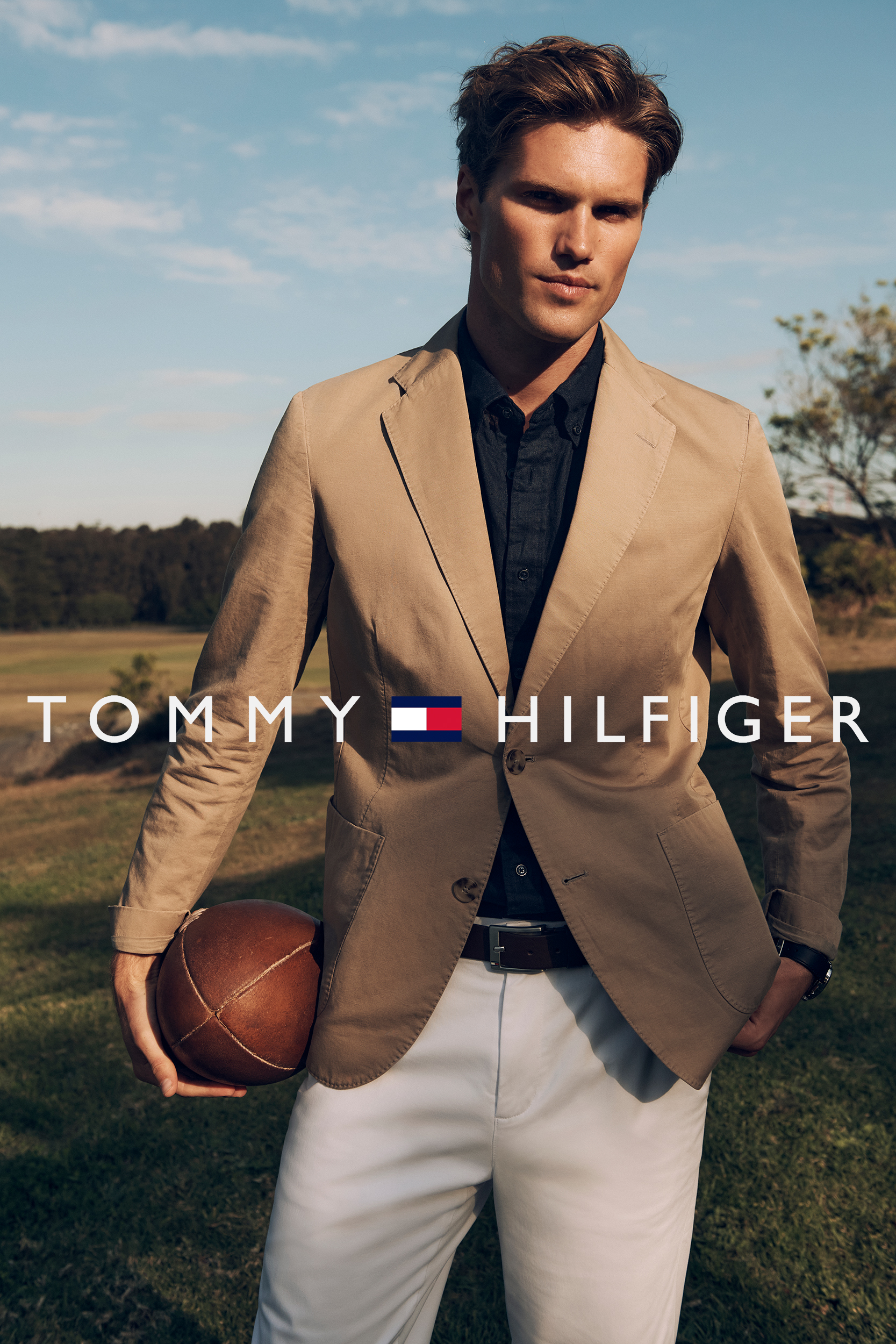 Fashion shot of an adult male in a tan suit jacket and white trousers, holding a football in an outdoor setting. Tommy Hilfiger logo.