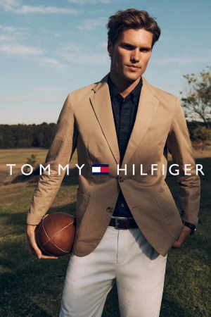 Fashion shot of an adult male in a tan suit jacket and white trousers, holding a football in an outdoor setting. Tommy Hilfiger logo.