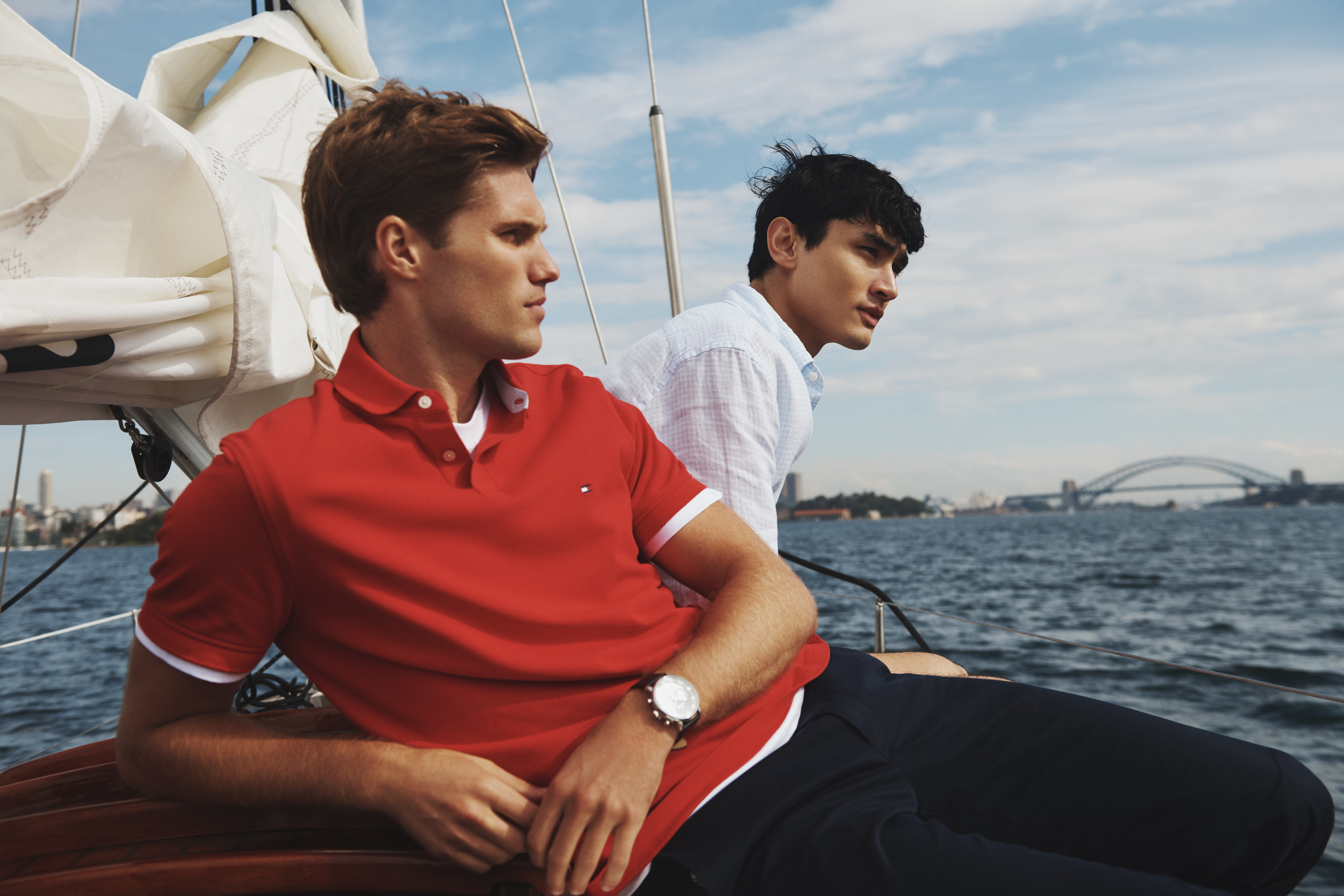 Two fair-skinned adult men in polo shirts sit on a sailboat with navy pants, with a partial view of a bridge over water in the background.