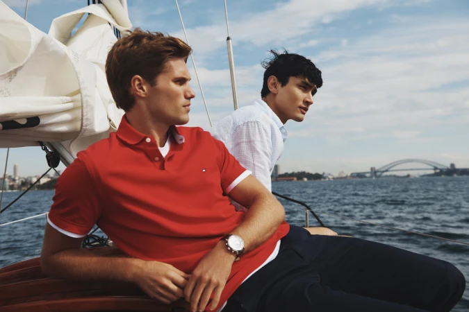Two fair-skinned adult men in polo shirts sit on a sailboat with navy pants, with a partial view of a bridge over water in the background.