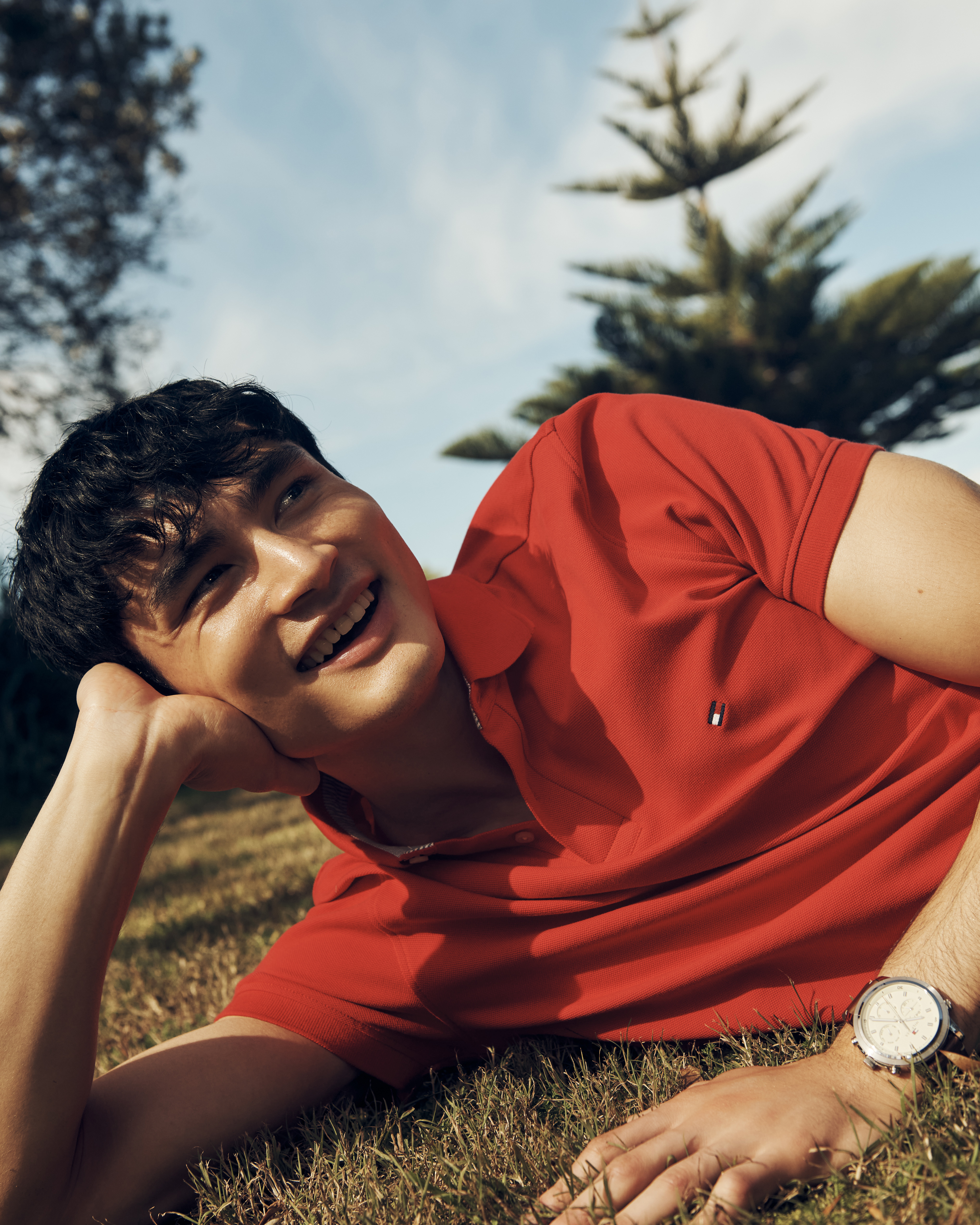 Smiling adult male model in red polo shirt, lying on grass in outdoor setting with blue sky and trees in the background.