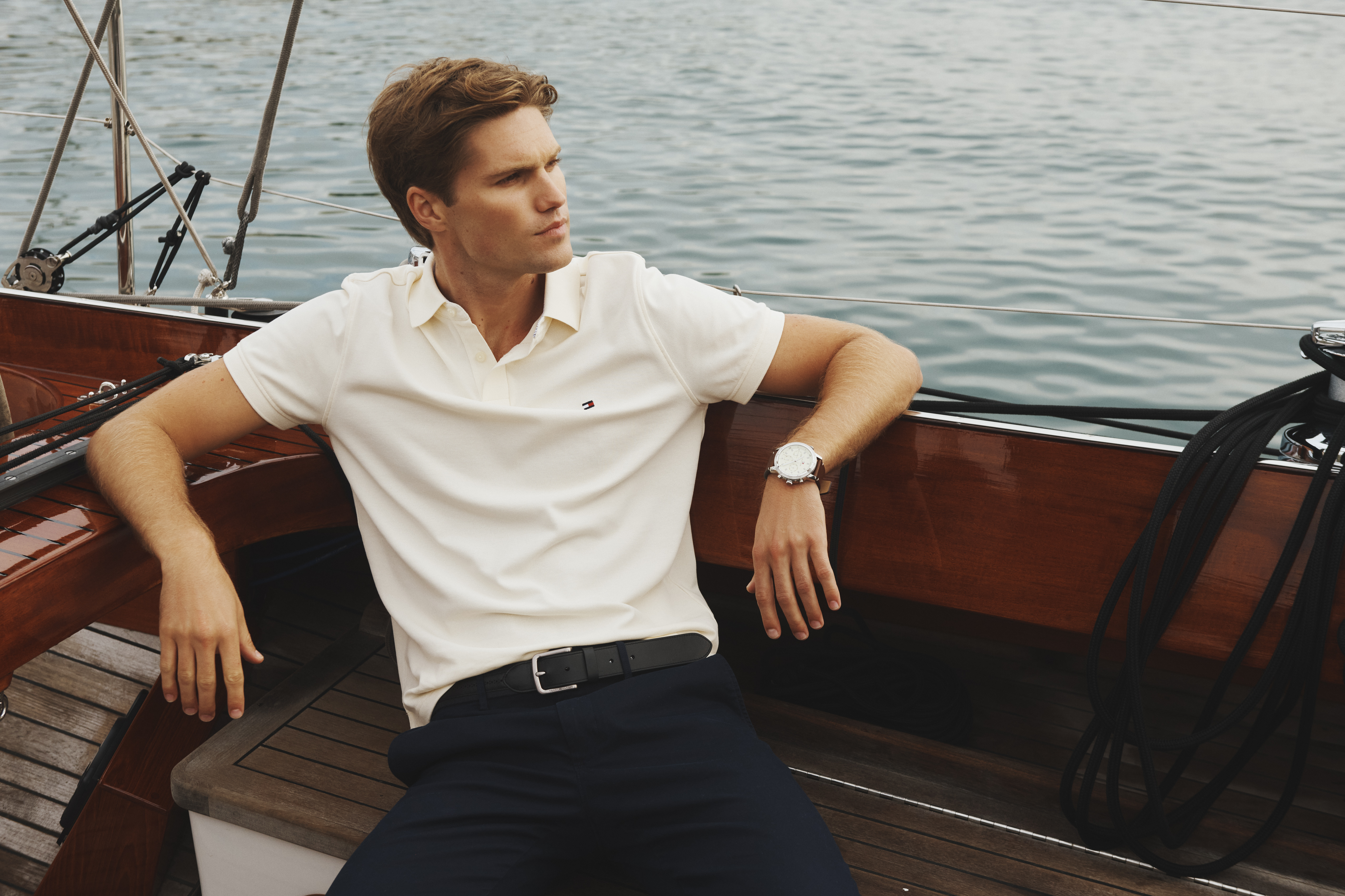 Handsome fair-skinned man with short brown hair, wearing a polo shirt and dark slacks, relaxes on a wooden sailboat in daylight.