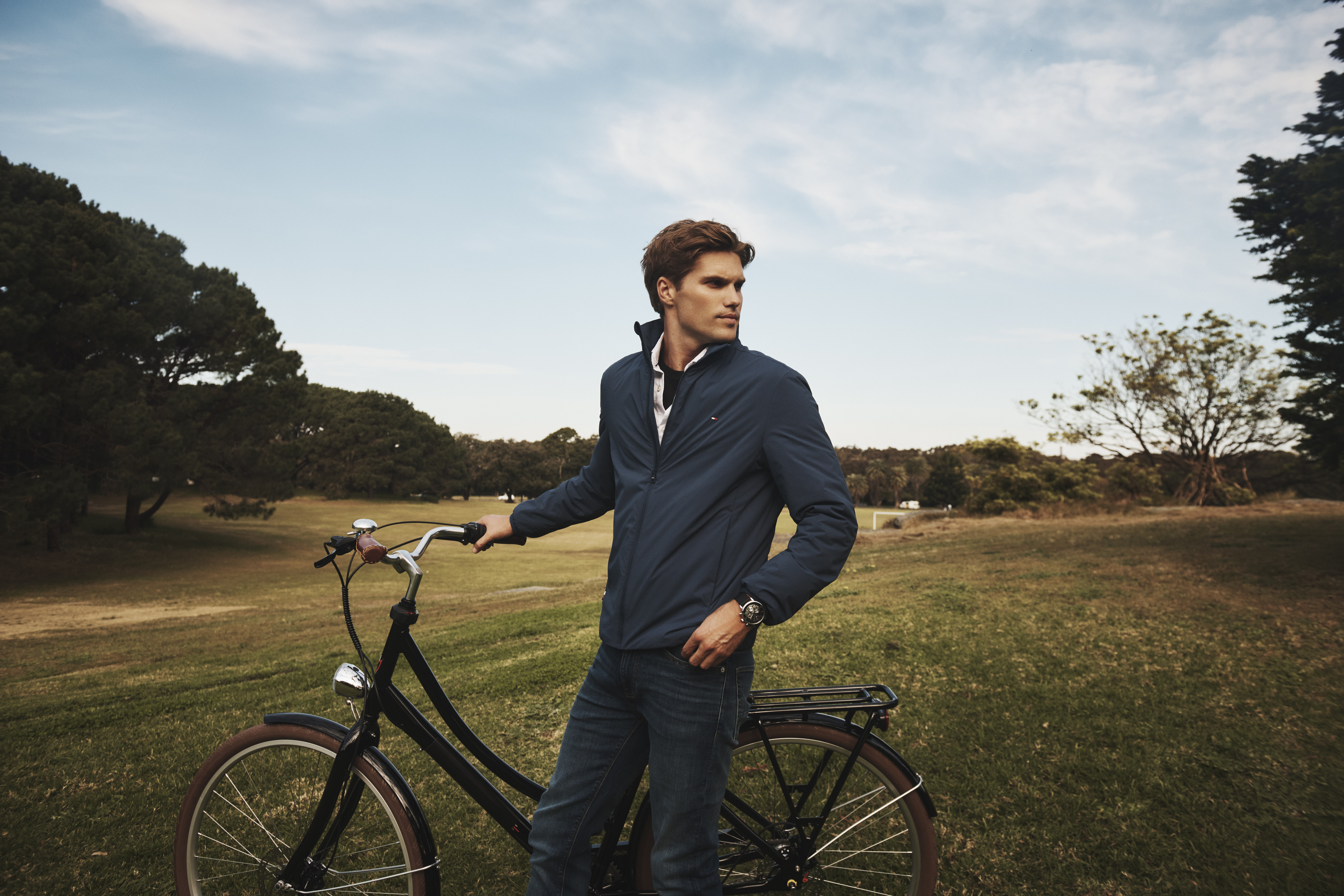 Adult male stands with a black bicycle on a grassy field under a bright sky, surrounded by trees, wearing a blue jacket and jeans.